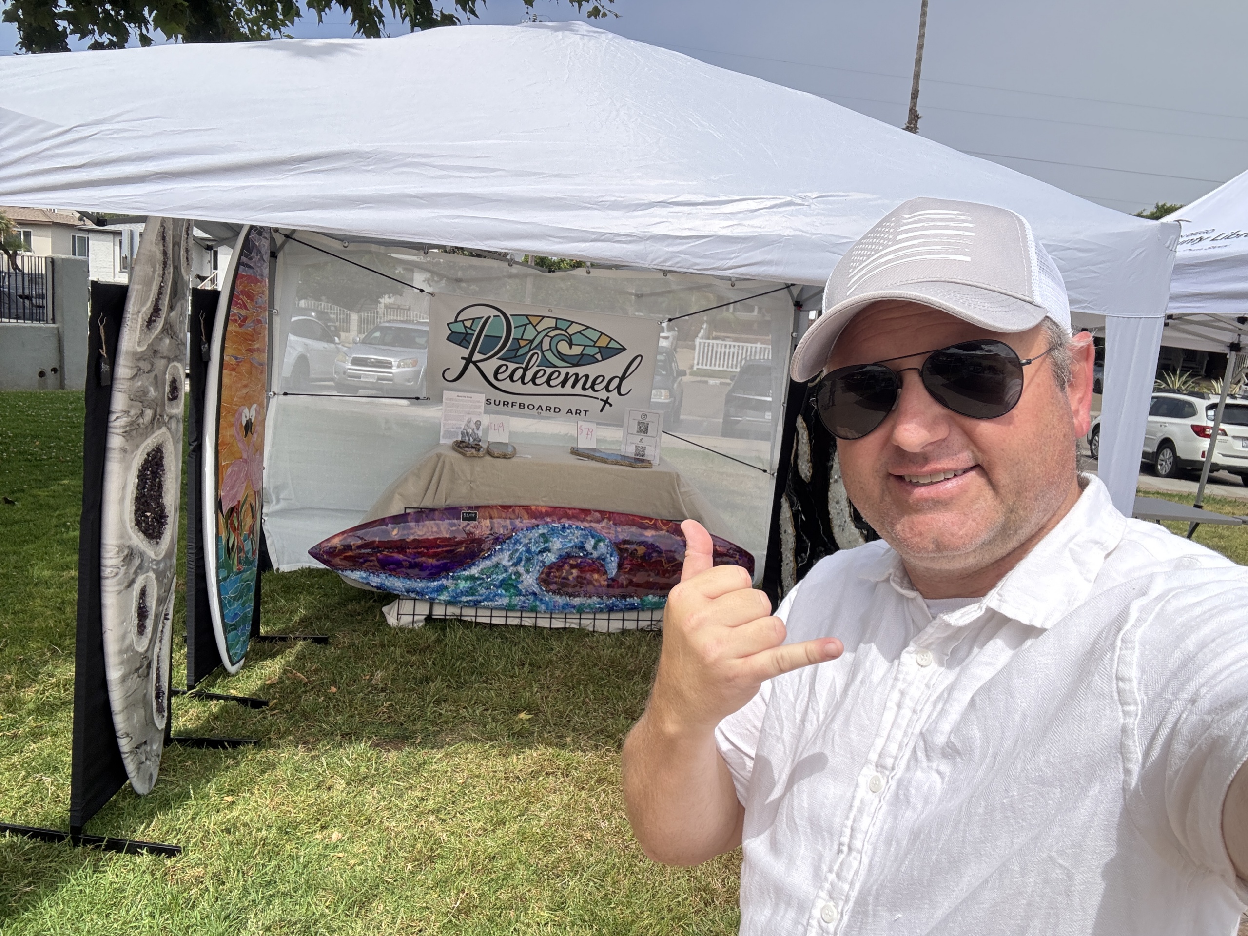 male in white shirt standing in front of art booth holding the shaka hand pose